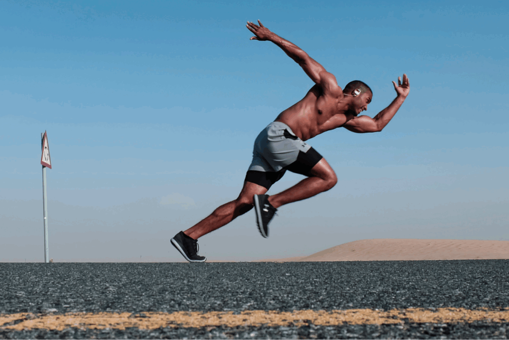 Topless man wearing grey and black shorts running on tarmac road with blue sky behind.