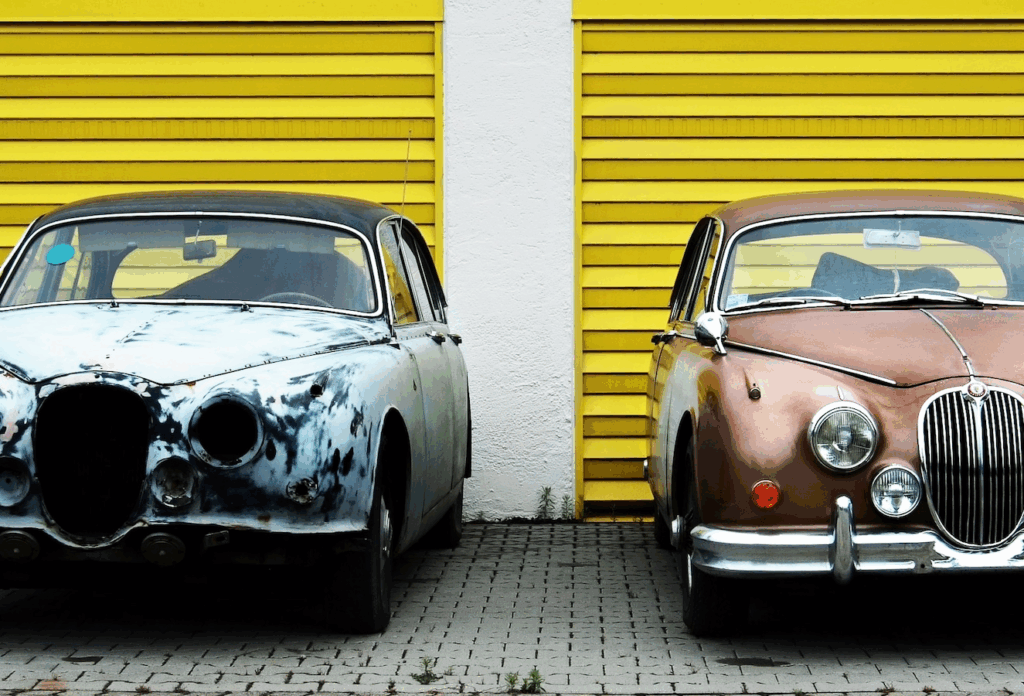 Two cars next to each other in front of two yellow garage doors. One car is rusty and the other is in good condition.